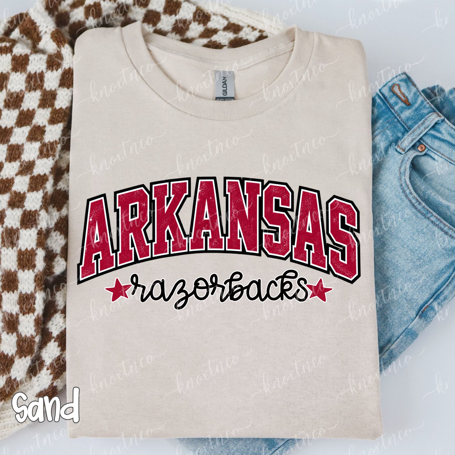 Sand unisex Arkansas Razorbacks game day t-shirt with vintage distressed crimson arch lettering and black script font, styled flat lay with denim jeans