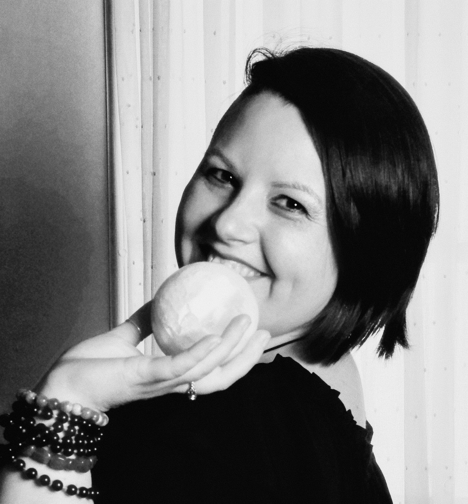 Photograph of Evylyn Rose in black and white. She is smiling with joy and holding a selenite sphere that appears like a small moon. Her wrist is adorned with several crystal bracelets. Photograph by Matanda Mondoa.
