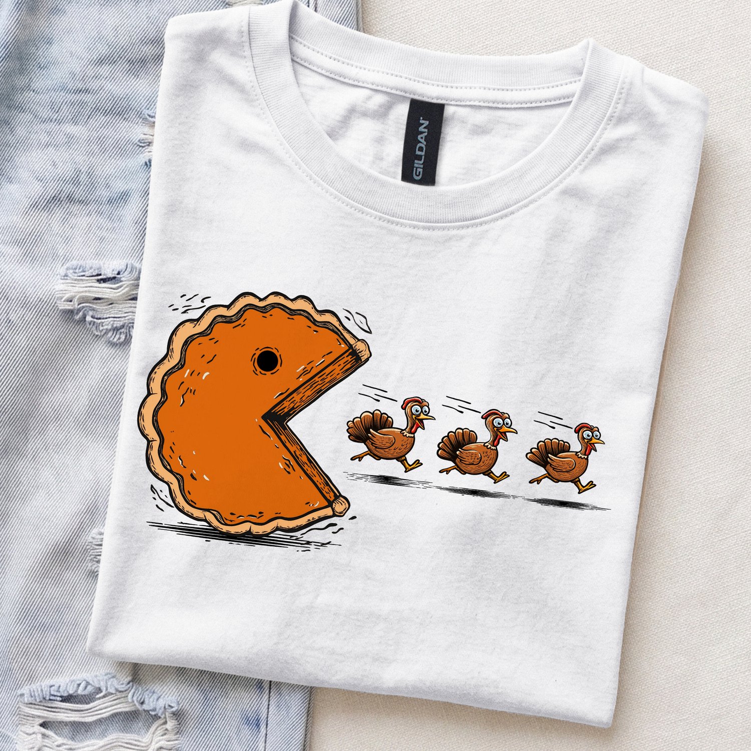 Flat lay mockup of a white Gildan Softstyle t-shirt folded next to distressed denim jeans, featuring a graphic print of a giant pumpkin pie chasing three running cartoon turkeys in a retro arcade video game style.
