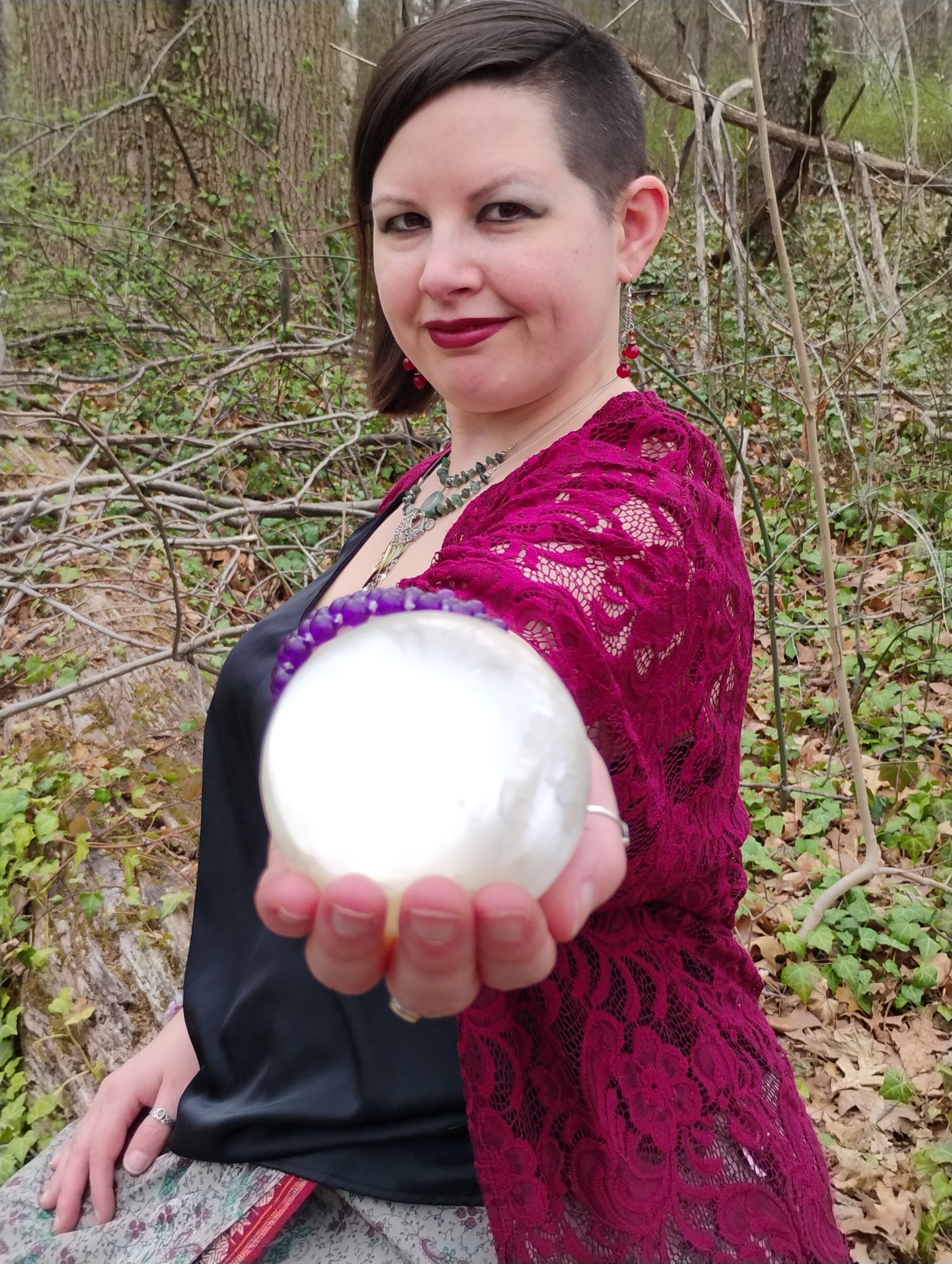 Photograph of Evylyn Rose looking toward the direction of the camera, left arm outstretched, holding a selenite crystal ball to the camera. Photograph by Ryan Hobbs, 2025