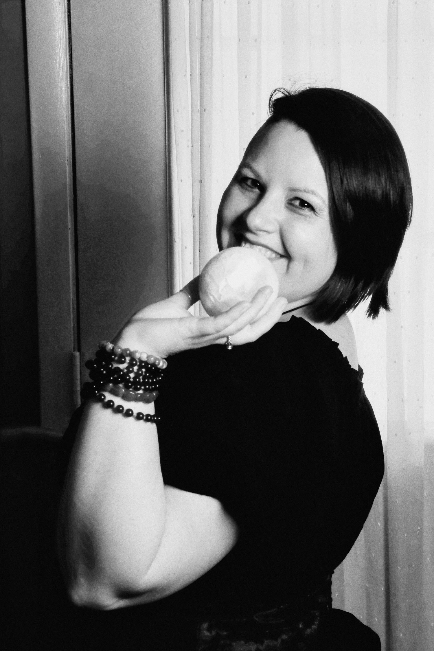 Photograph of Evylyn Rose in black and white. She is smiling with joy and holding a selenite sphere that appears like a small moon. Her wrist is adorned with several crystal bracelets. Photograph by Matanda Mondoa.