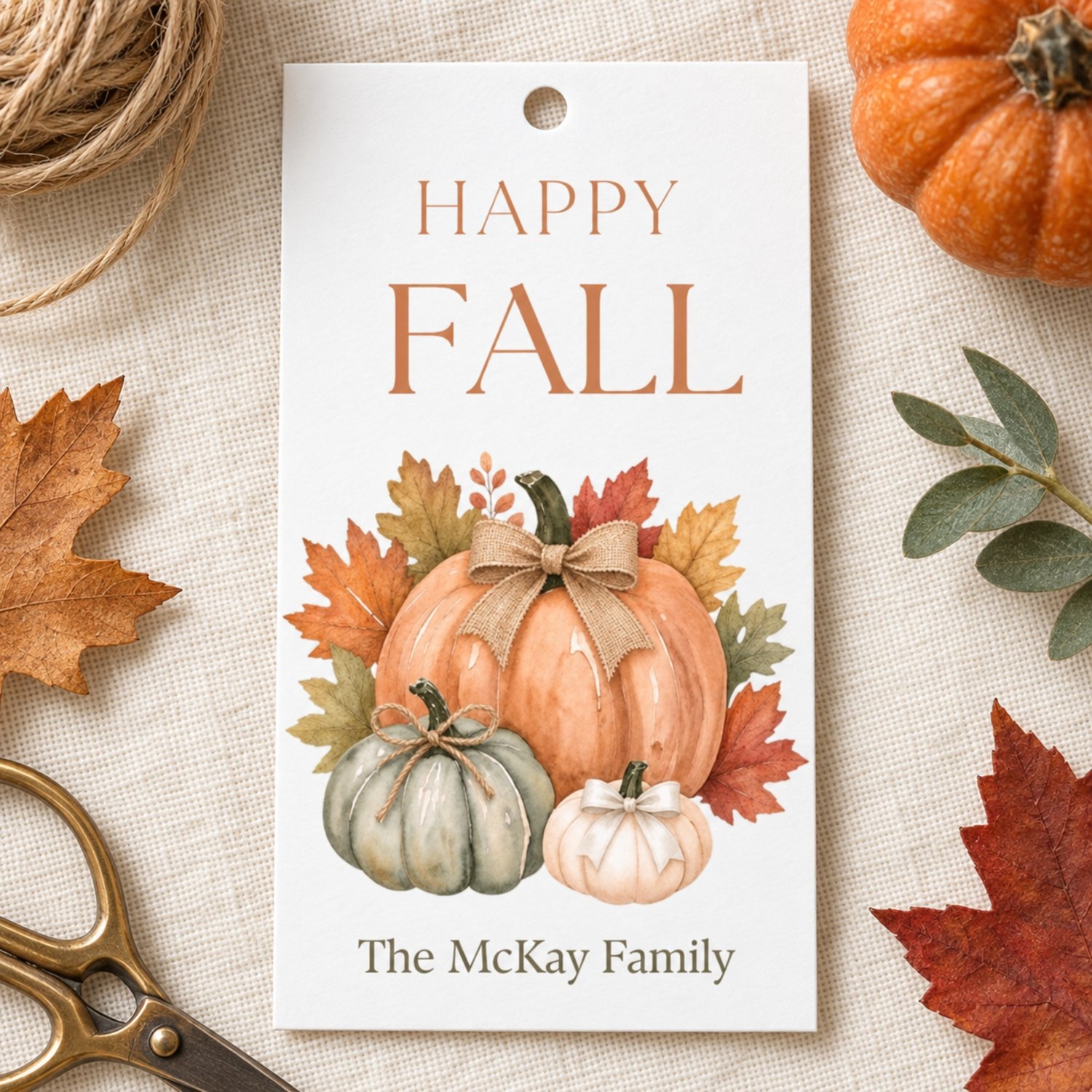 A rectangular white gift tag temaplate featuring the text HAPPY FALL in brown letters at the top and editable text at the bottom. The center displays a watercolor illustration of three pumpkins in orange, green, and white with autumn leaves.