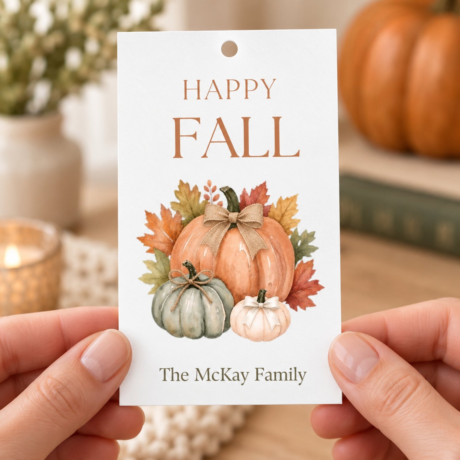 A rectangular white gift tag temaplate featuring the text HAPPY FALL in brown letters at the top and editable text at the bottom. The center displays a watercolor illustration of three pumpkins in orange, green, and white with autumn leaves.