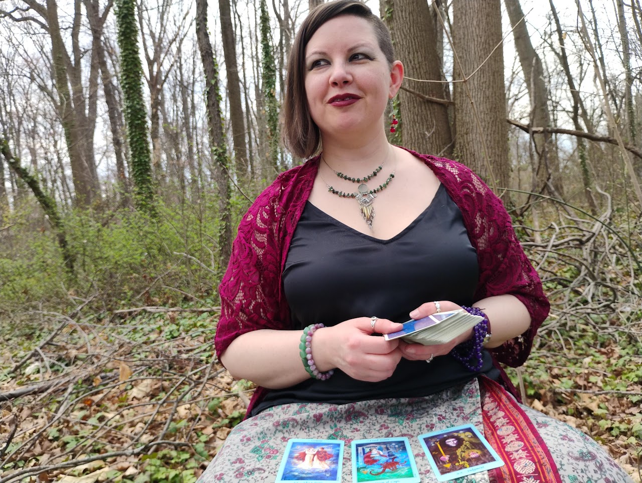 Photograph of Evylyn Rose looking out into the woods where she is sitting. She has three Tarot cards in a spread in front of her. Photograph by Ryan Hobbs, 2025