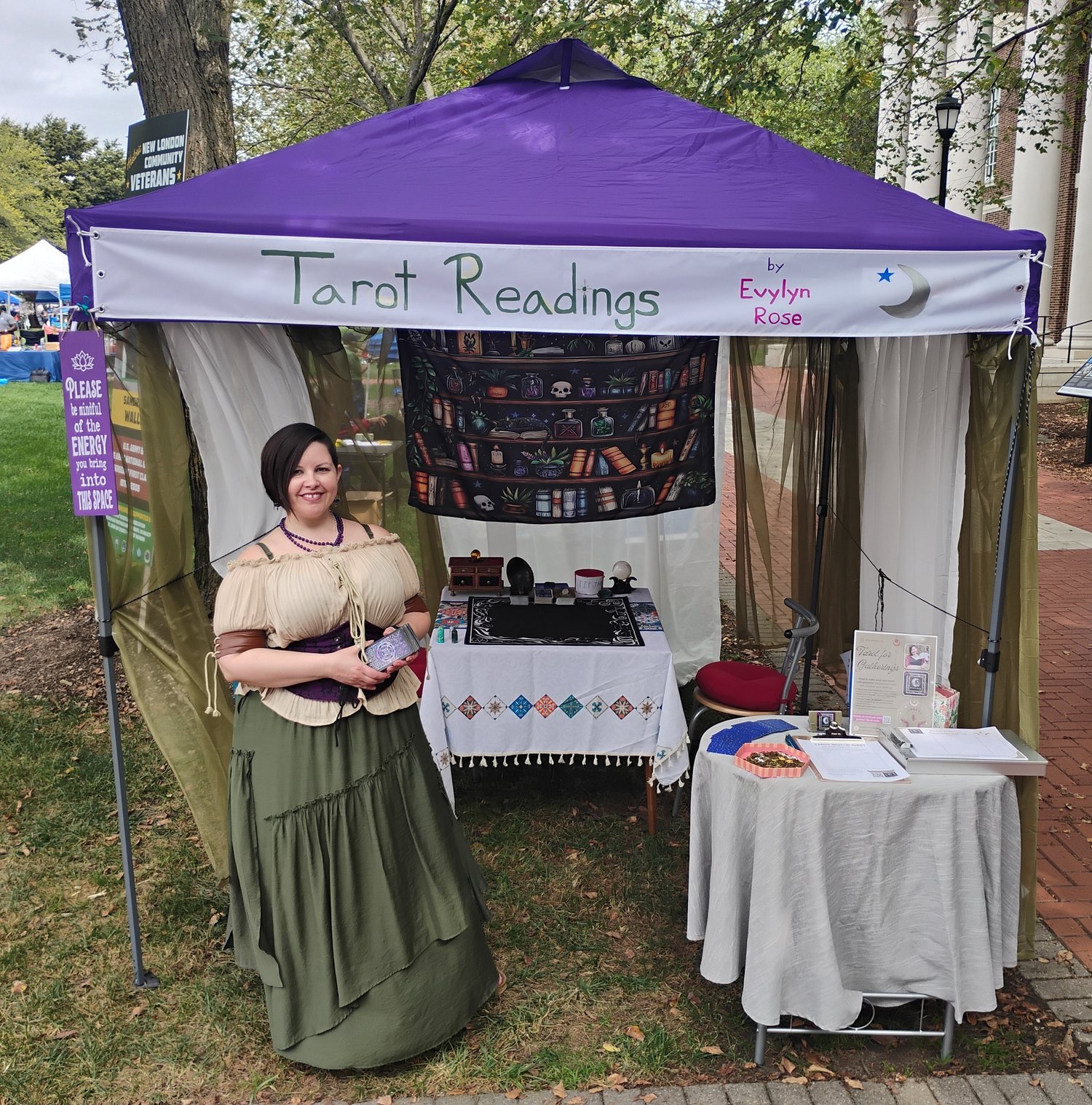 Photograph of Evylyn Rose holding a Tarot deck at her Tarot Readings booth for the 2025 Newark Community Day in Delaware.