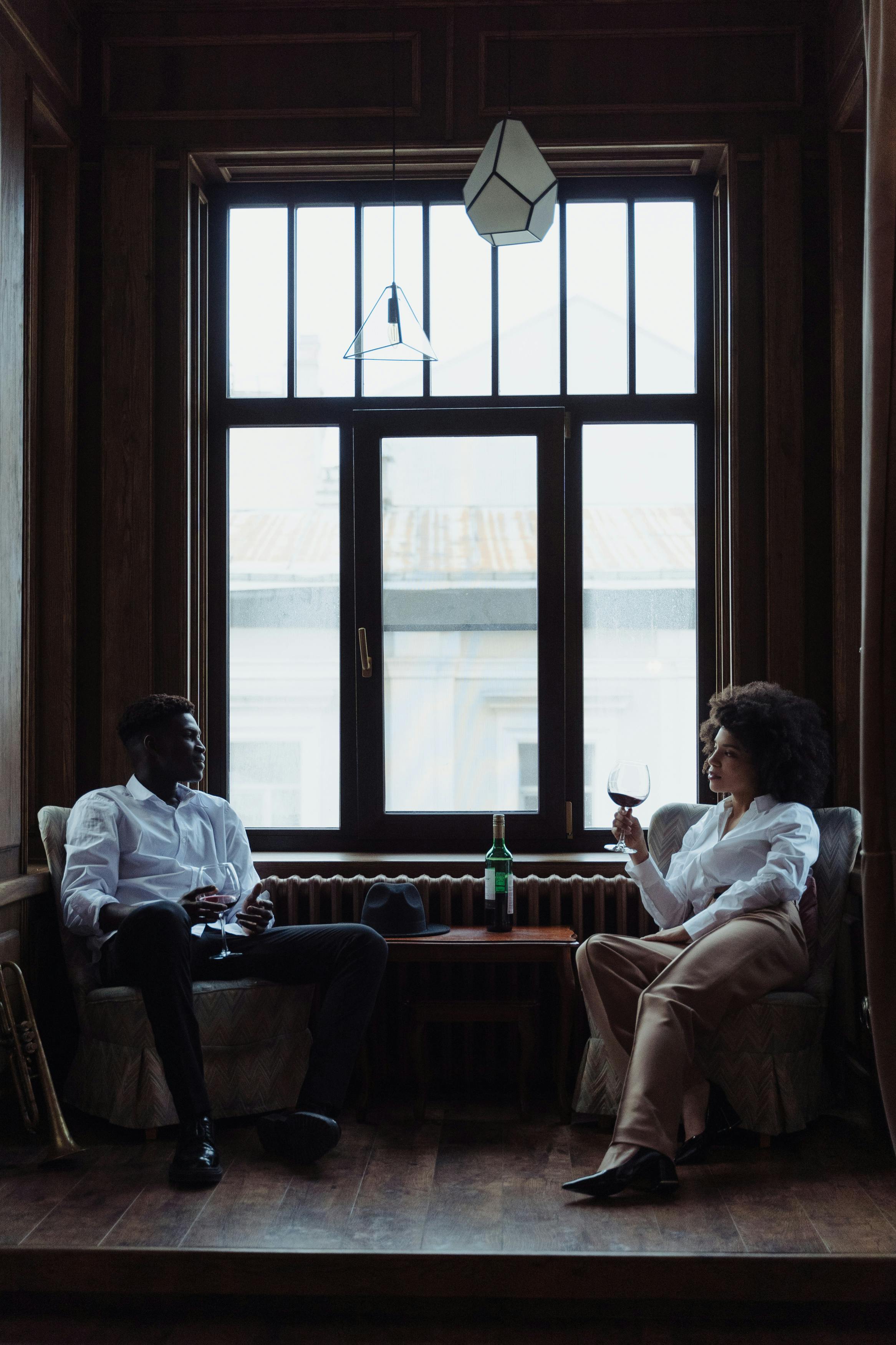 black -adult-male-with-white-shirt -seating by a windowm witha-lady dressed in neige pants and a white dress shirt both are holding a glass of wine seated across each other