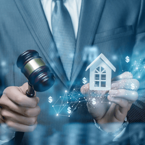 auctioneer in grey suit with grey tie holding gavel and small white toy home with dollar signs