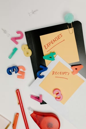 white background with black notebook, red pen, and red white-out dispenser with magnetic numbers and sticky note labels with expenses and receipts