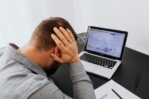 black desk with laptop and white male in grey shirt with head in hands looking at disappointing financial data