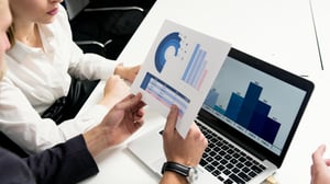 a woman in a white shirt and a man in a black suit jacket are looking at data on a print out and a laptop.