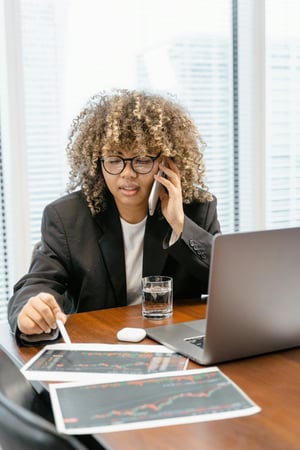 black woman in black blazer is sitting in office and analyzing charts and graphs while speaking on a cell phone