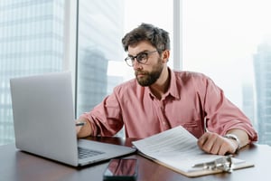 White male with beard and glasses in pink shirt is sitting in front of laptop and flipping through a clipboard