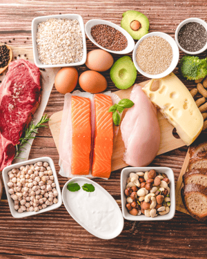 A variety of high-protein and healthy fat foods, including eggs, salmon, chicken breast, nuts, avocados, and olive oil, arranged on a clean surface to highlight nutritious meal choices for muscle growth and weight management.