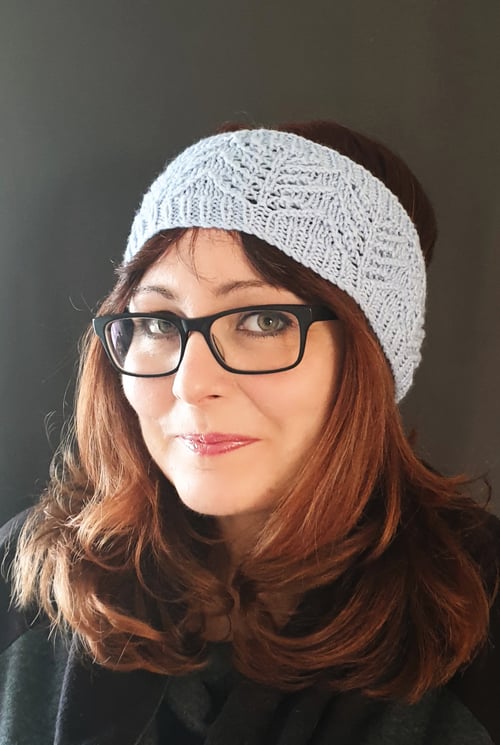 A dark haired lady wearing glasses. She has a knitted ice blue headband and a dark background.