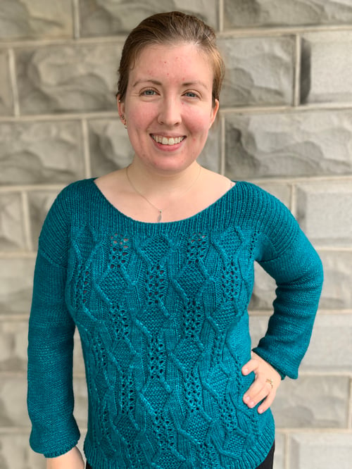 Ruth stands, smiling, wearing her Arkenstone Pullover design. It is a teal, long sleeved sweater with cables and lace down the front.