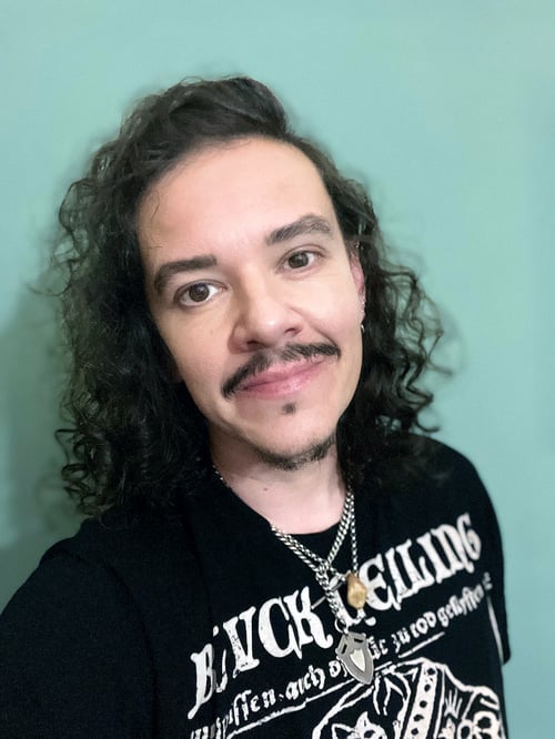 Author Die Booth, a white middle aged man with long dark curly hair, dark eyes and dark facial hair, wearing a black 'Blvck Ceiling' tshirt and chain necklaces, standing against a pale green background
