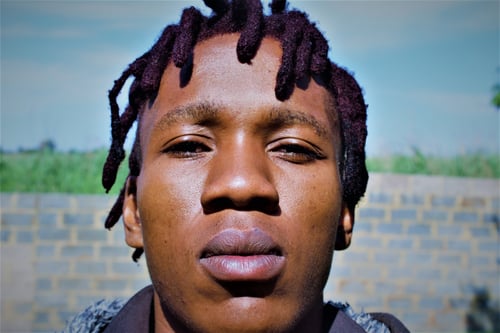 Obakeng Masego Mooketsi, an African male with a light brown complexion and distinctive maroon-colored dreadlocks, stands confidently against a vibrant blue skyline background. As the owner and founder of Black Goat Inc., his professional and charismatic presence reflects leadership and innovation.