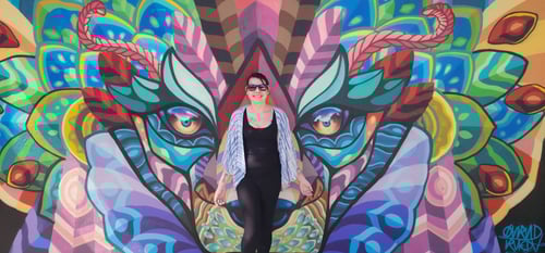 Smiling woman with long brown hair standing in front of a colorful mural, friendly and creative small business owner, maker of printable SEL resources, teacher tools, lunchbox notes, and custom crafts for kids and families
