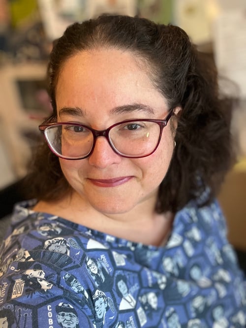 Photo of a white woman wearing magenta eyeglasses with dark brown hair. She's wearing a blue dress.