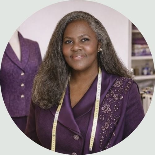 Professional portrait of Carolyn, founder of The Academy of Sewing, wearing a purple jacket with a measuring tape around her neck in her sewing studio.