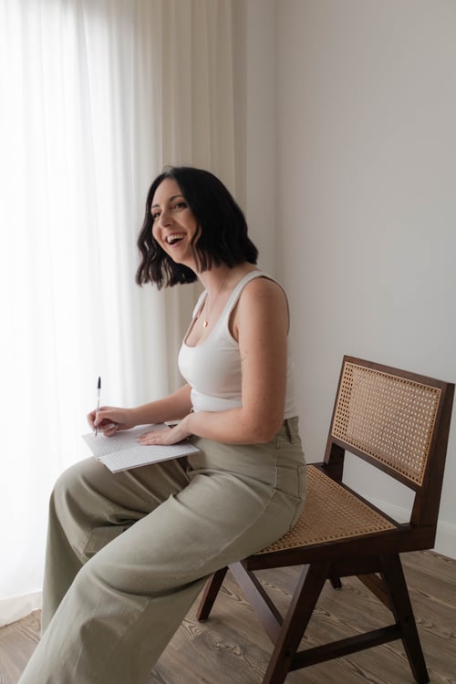Dr. Lauren Cooper, PhD English Literature tutor and self-reflection workshop facilitator, sitting in a cane chair in a minimal bright space, writing in a notebook and laughing — representing a gentle, joyful approach to journaling and literary self-study