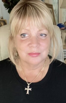 Close‑up portrait of a woman with blonde hair, wearing a black shirt and gold cross necklace, photographed indoors.”