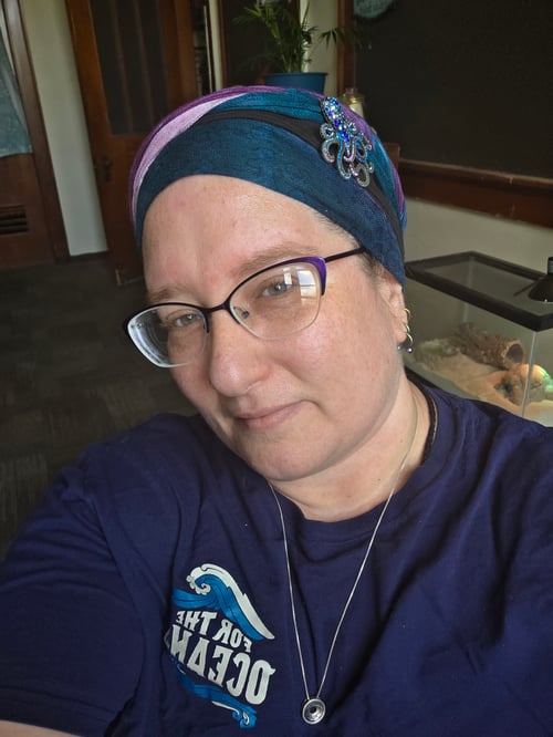 head, neck, and chest of a white person wearing a dark blue shirt, matching headwrap with an octopus brooch for decoration, and eyeglasses