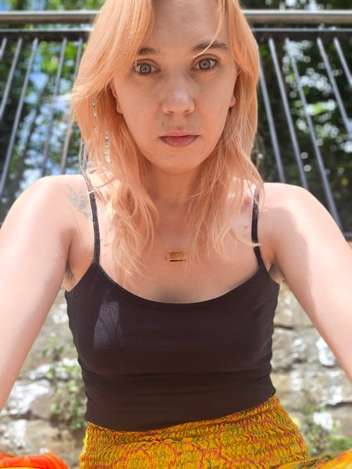 woman sitting in a park with a fence in the background, light blonde strawberry blonde hair, blue eyes, black strappy top tshirt vest top with yellow casual spiritual trousers with a brick wall behind and little bits of sky visible