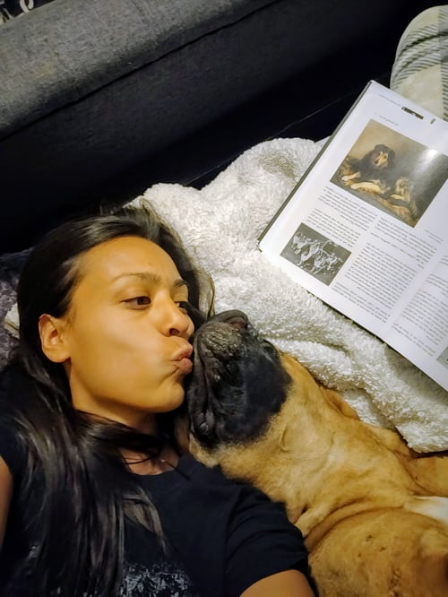 A close-up photograph of a woman—a cynologist and founder of Project Guerrero Bullmastiff—relaxing indoors with her working Bullmastiff. She is leaning in to kiss the dog while lying next to an open book featuring historical canine illustrations and text on working dogs. The setup highlights the deep bond, companionship, and commitment to the functional preservation and study of the historic Nightdog type.