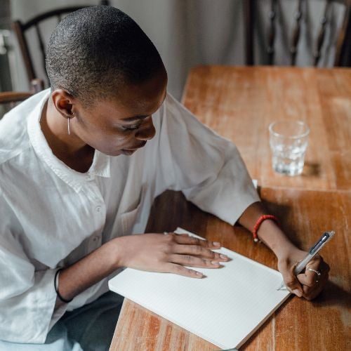 Woman writing with her left hand in a notebook while seated at a wood table