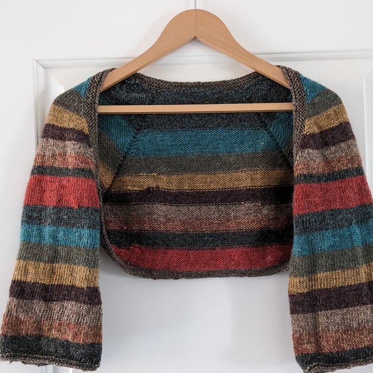 stripy knitted shrug garment hanging on a wooden coathanger, the colours are autumnal