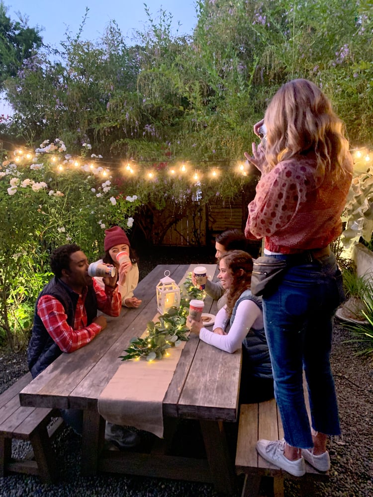 Four people sit at a rustic wooden table outdoors, surrounded by greenery and string lights. Another person stands taking a photo of them. They all hold drinks, and a lantern with a garland decorates the table. Its twilight, creating a cozy atmosphere.