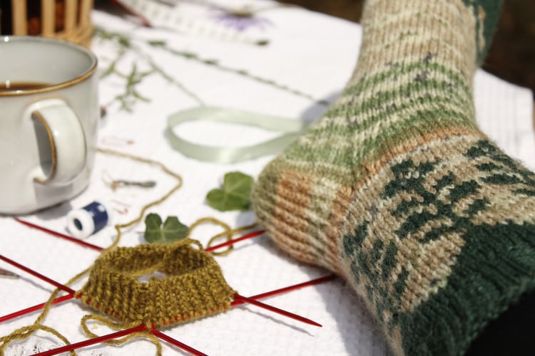 A colorwork sock featuring a flower motif in earthy tones of green and beige, resting on a table. Someone is knitting a new sock in olive green and drinking coffee. A relaxing day, knitting in the garden in a sunny Provence, France.