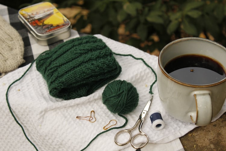 A forest green sock being knit while drinking a cup of coffee, in the garden. A cozy and crafty time in Provence, France.