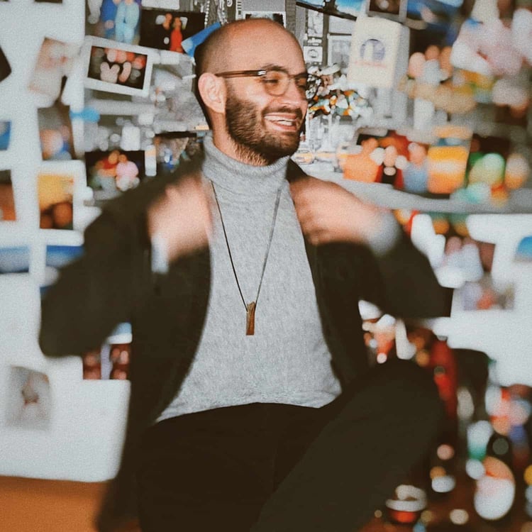 Selbstbewusster Mann mit gepflegter Glatze und Vollbart trägt Rollkragenpullover, lächelt authentisch vor Fotowand im kreativen Ambiente.