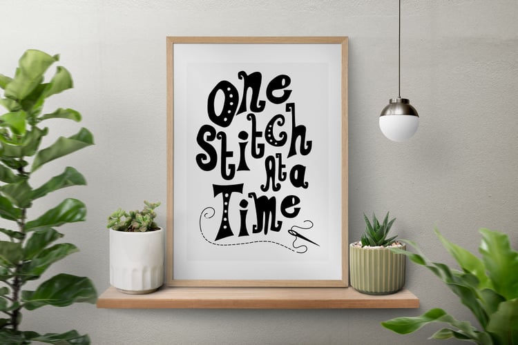 A downloadable print with a hand lettered design which says ‘one stitch at a time’ in black.