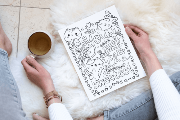A person colors a "You are the sweetest" illustration with cats and flowers, holding a cup of coffee, seated on a cozy fuzzy rug.