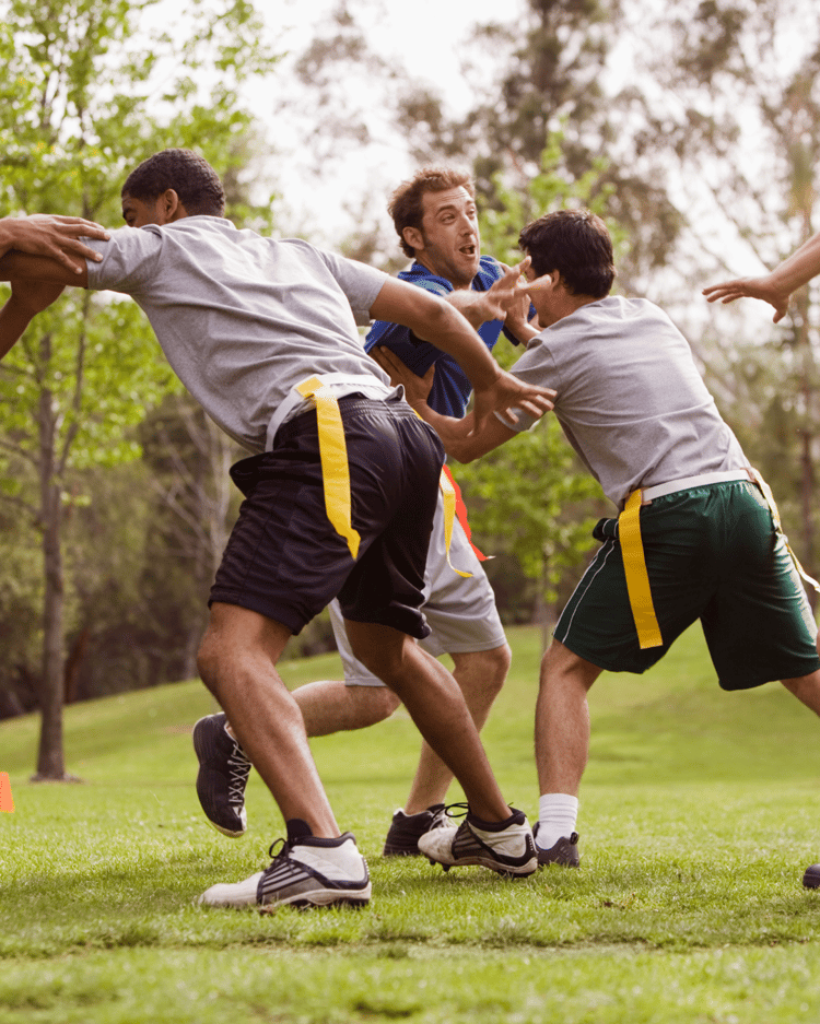 A group of men over 40 playing flag football on a grassy field, displaying teamwork, determination, and camaraderie. The players are actively engaged, running, passing, and strategizing, symbolizing the importance of sports in building fitness, social connections, and mental toughness