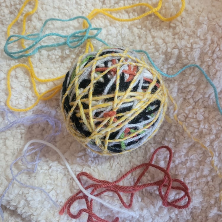 A ball of colorful scrap yarns tied together sits on a beige carpet.