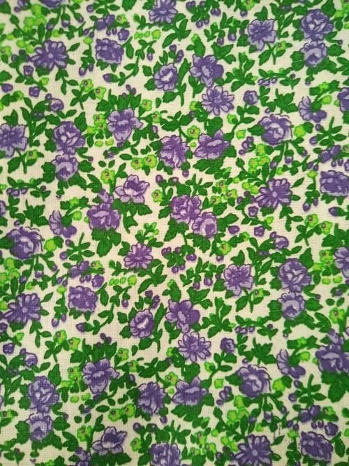 MDG 100% cotton calico fabric with purple and lilac flowers, green leaves, and stems on an off-white background, shown as a 3-yard unwashed cut off the bolt
