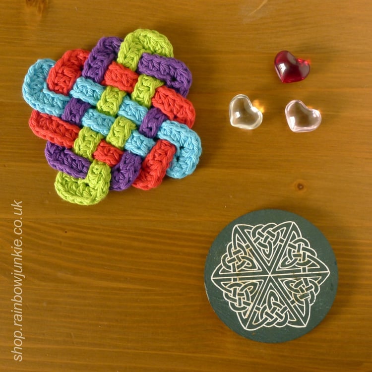 Four part Celtic plaitwork coaster made in worsted weight yarn in red, blue, green and purple on a table layout.