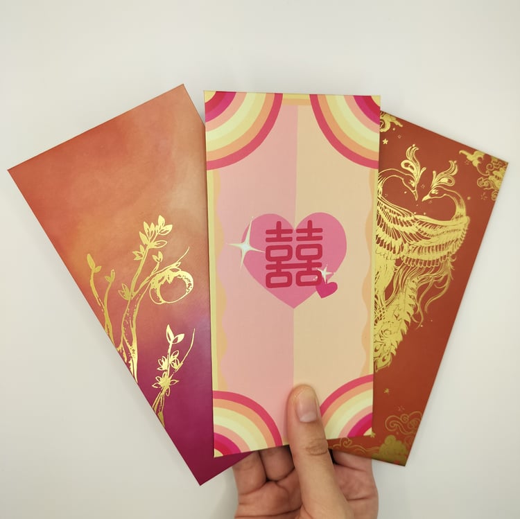 Hand holding three Chinese New Year money envelopes (hongbao) - an orange-to-red card with gold botanical foil, a rainbow striped card with pink double happiness heart, and a red card with gold dragon design.