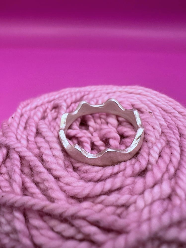 Silver ring with wavy, scalloped edges nestled in coiled pink yarn against a vibrant magenta background.