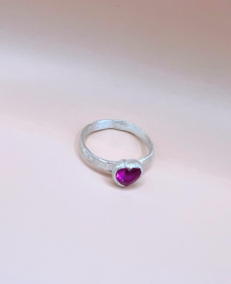 Silver ring with a textured band featuring a heart-shaped pink ruby in a bezel setting against a neutral beige background.