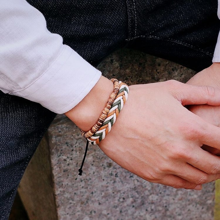 Mans wrist showcasing a 2-piece Bohemian bracelet set: includes a natural wood rondelle bead bracelet and a wide flat-braided cord bracelet in neutral friendship colors (olive green, cream, tan). Styled casually with a white shirt and dark denim jeans ou