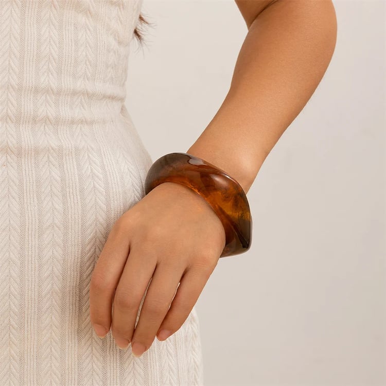 Lifestyle image of a woman wearing a stylish chunky wide cuff bangle made of dark brown tortoiseshell resin. The statement bracelet's unique organic square shape is shown on her wrist, styled with a white ribbed halter top. Modern fashion accessory.