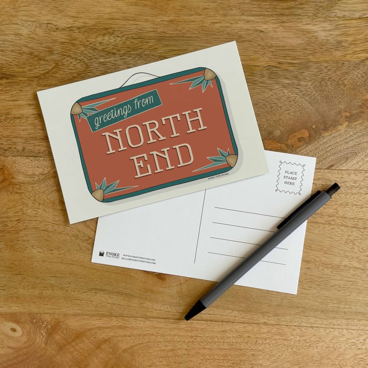 The front and back sides of a postcard with a pen placed on top. The front features a vintage suitcase illustration and the back shows the address lines and stamp indicator.