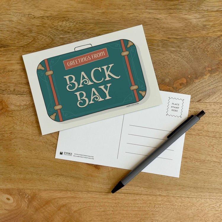 The front and back sides of a postcard with a pen placed on top. The front features a vintage suitcase illustration and the back shows the address lines and stamp indicator.