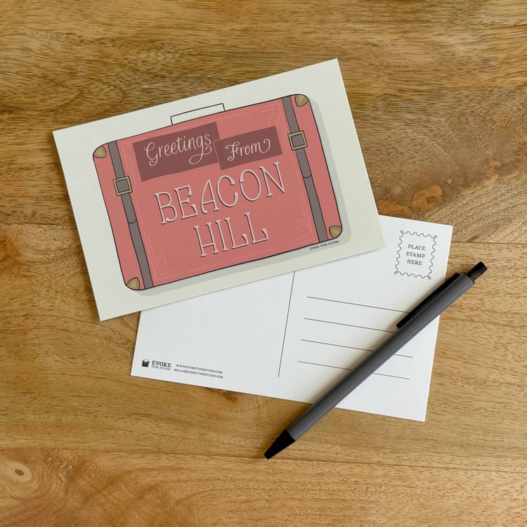 The front and back sides of a postcard with a pen placed on top. The front features a vintage suitcase illustration and the back shows the address lines and stamp indicator.