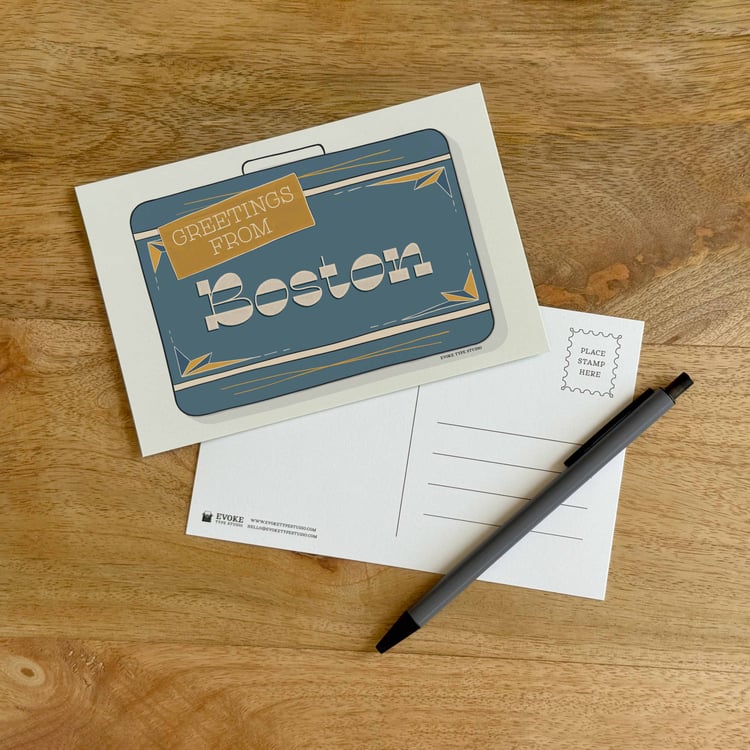 The front and back sides of a postcard with a pen placed on top. The front features a vintage suitcase illustration and the back shows the address lines and stamp indicator.